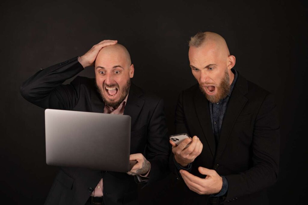 Two traders reacting emotionally to sudden market movements on a laptop and smartphone, illustrating how stubbornness and emotional attachment can lead to poor trading decisions instead of objective analysis.