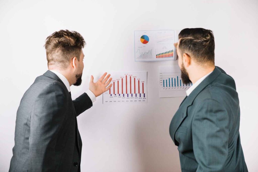 Two traders analyzing market charts and performance data, illustrating the difference between predicting market movements and reacting to changing market conditions in trading.