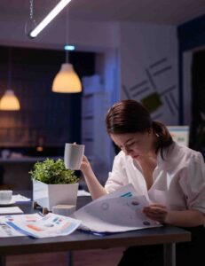 A focused woman sitting at a desk in a dimly lit office at night, holding a white coffee mug while carefully reviewing financial documents and charts, illustrating a disciplined, analytical approach to trading and managing investment positions.