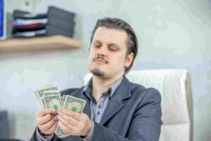 A man in a grey suit jacket sitting in an office chair, looking down with a skeptical or uncertain expression as he counts a small fan of one-dollar bills, illustrating the concept of a trader securing minor, premature gains instead of letting a winning investment grow.